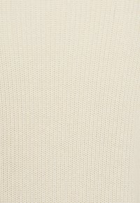 Crèmewit gebreide stof met een fijn, gestructureerd patroon en geribbelde afwerking. Het materiaal lijkt zacht en ademend.