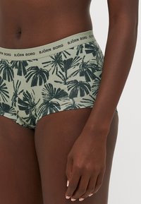 Green patterned underwear with a wide elastic waistband labeled "BJÖRN BORG." Features dark palm leaf designs, short leg cuts, and a soft texture.