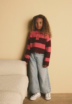 Sweter w paski w kolorze burgundowym i różowym z kołnierzykiem i guzikami, zestawiony z luźnymi, jasnymi dżinsami i białymi sneakersami. W pobliżu teksturowana biała sofa.