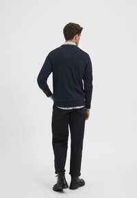 Suéter de punto azul marino con cuello redondo; llevado encima de una camisa de cuadros. Los pantalones negros y las botas negras completan el atuendo.