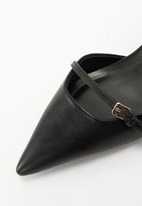 Chaussure noire en cuir à bout pointu avec une sangle délicate autour de la cheville et un accent de boucle en or, présentant une texture lisse et un design minimaliste.