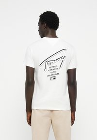 T-shirt en coton blanc avec un design textuel graphique noir au dos, comprenant des noms de villes et une icône de cassette. Manches courtes, coupe standard.