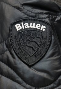 Μαύρο υφασμάτινο καπιτονέ με ένα μαύρο και λευκό κεντητό έμβλημα σε σχήμα ασπίδας Blauer που παρουσιάζει έναν στυλιζαρισμένο αετό και κείμενο.