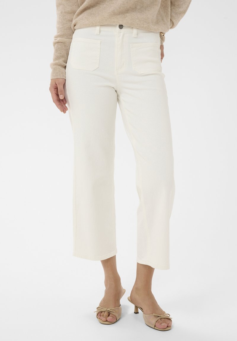 Femme portant un jean droit coupé écru, un pull beige et des sandales à talons kitten ouvertes de couleur nude, debout sur un fond blanc.