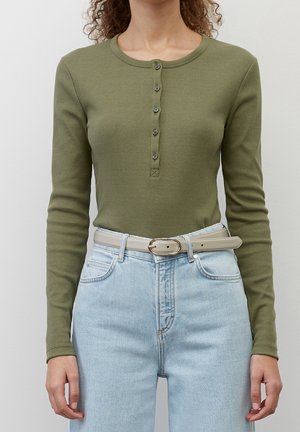 Frau trägt ein olivgrünes, geripptes Langarm-Henleyshirt, das in hellblaue, hochtaillierte Jeans mit einem beigen Gürtel gesteckt ist.