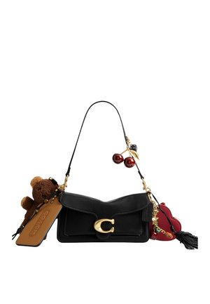 Sac bandoulière en cuir noir avec fermoir doré, décoré d’un ours en peluche, d’une étiquette beige, d’un pendentif cœur rouge, d’ornements cerise et dés, et d’un pompon noir.