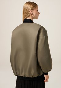 Giacca bomber verde oliva realizzata in materiale simile alla satin, con colletto e polsini a costine neri, dal taglio rilassato e oversize.
