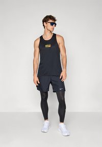 Juoda sportinė liemenė su „BERLIN RUN“ logotipu, derinama su tamsiais šortais ir juodomis kompresinėmis tamprėmis. Baltos sportinės avalynės ir saulės akiniai.