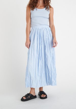 Kobieta ubrana w jasnoniebieską, bezrękawnikową bluzkę i długą pomarszczoną spódnicę, w czarnych sandałach na płaskiej podeszwie, stojąca na tle jednolitego białego tła.