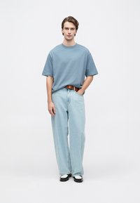 Jauns vīrietis stāv, ģērbies brīvā zilo t-kreklā, gaiši zilos plašos džinsos ar brūnu jostu un melnbaltās kurpēs, rokas atbrīvotas.