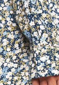 Chemise à motifs floraux en bleu, blanc et jaune. Tissu en coton, avec de petites fleurs et des motifs feuillus, détail de poignets à boutons.