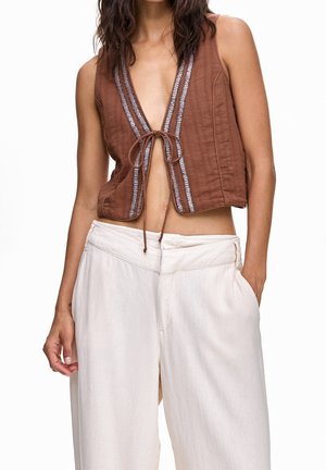 Woman wearing sleeveless brown cropped vest with front tie and high-waist loose white pants, one hand in pocket.