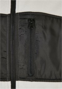 Black zippered pocket on a beige fleece jacket, featuring a contrasting black nylon lining and smooth texture, with visible stitching details.