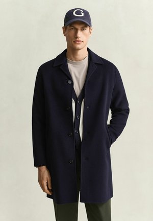 Mann trägt marineblauen Mantel, graues Hemd, marineblauen Cardigan und marineblaue Kappe mit Buchstabe G, steht vor einfachem hellem Hintergrund.