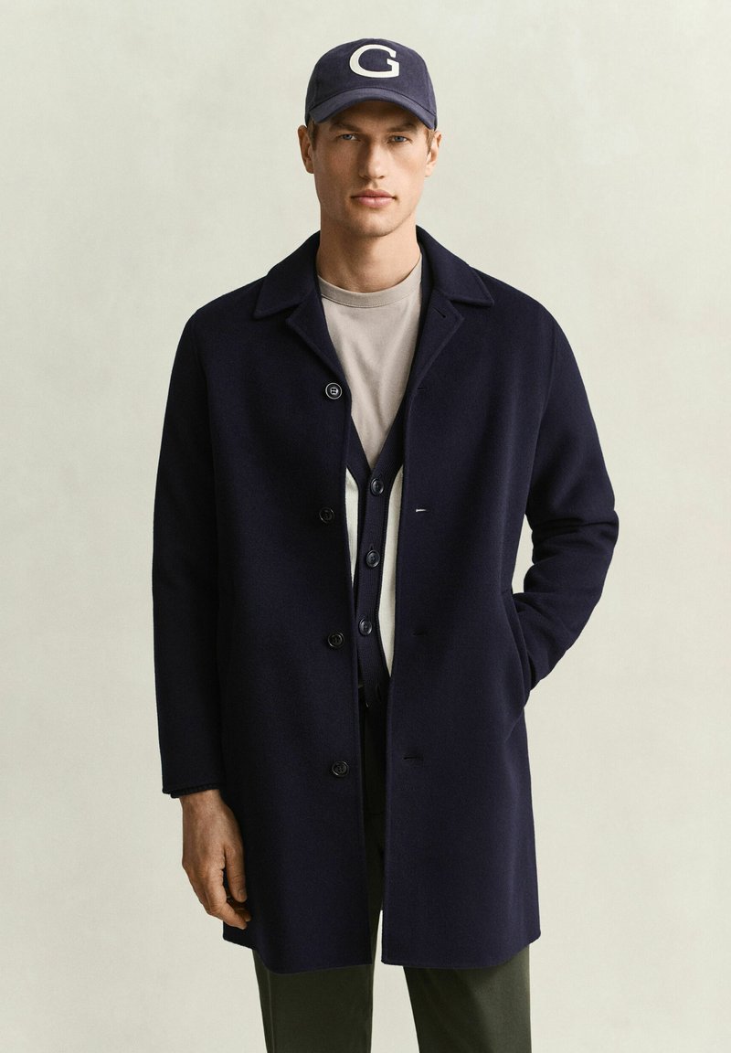 Homme portant un manteau bleu marine, une chemise grise, un cardigan bleu marine et une casquette bleu marine avec la lettre G, debout devant un fond clair uni.