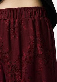 Bordo mežģīņu pidžamas bikses ar elastīgu vidukli, rotātas ar ziedu rakstiem un mīkstu tekstūru, daļēji redzamas no jostas līdz lejai.