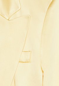 Zbliżenie na jasnopomarańczowy satynowy rewer i kieszeń piersiową z widocznym przeszyciem oraz gładką teksturą tkaniny.