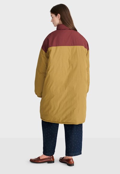 Femme aux longs cheveux bruns portant une longue veste bicolore moutarde et bordeaux, un pantalon bleu foncé, des chaussettes rayées et des mocassins marron, tourné vers la gauche.