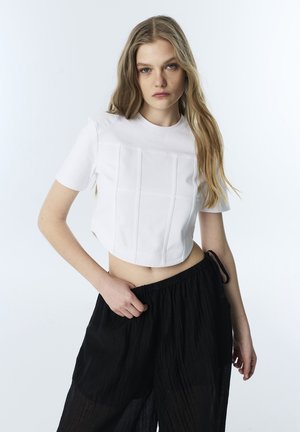 Mladá žena s dlouhými blond vlasy, která nosí bílý krátký top s krátkým rukávem a volné černé kalhoty na světlém pozadí.