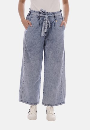 Persona che indossa pantaloni di jeans azzurri a gamba larga con cintura annodata in vita, mani in tasca e sneaker bianche con strisce nere.