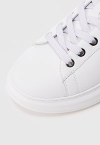 Bijela tenisica s glatkim gornjim dijelom od sintetičkog materijala, okruglim vrhom, ravnim bijelim potplatom i svijetloplavim vezicama provučenim kroz crne mrežice za vezanje.