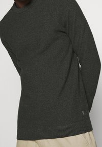 Pull en tricot vert foncé avec un col rond, des poignets et un ourlet côtelés, présentant une texture subtile et un petit logo en accent en bas.