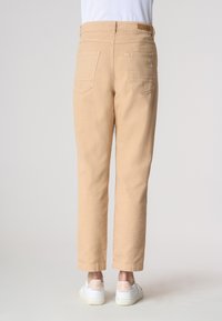 Pantaloni beige con taglio alla caviglia, indossati con sneaker bianche; la vista mostra il retro, evidenziando le tasche e il design della vita.