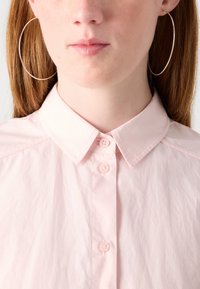 Gaiši rozā krekls ar uzvadām un apkakli, kuram ir divi pogas pie kakla. Lielas sudraba riņķa auskaru tuvplāns. Gluda tekstūra.