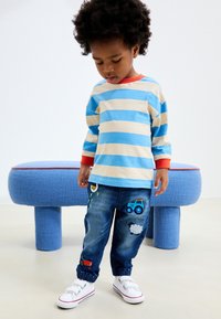 Blå og beige stribet langærmet skjorte, denim joggers med broderede detaljer, hvide sneakers og en blå tekstureret bænk i baggrunden.