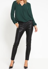 Blusa verde a V con maniche lunghe abbinata a pantaloni skinny neri in pelle ecologica e tacchi a punta neri, creando una silhouette elegante.
