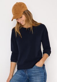 Marineblauer Sweatshirt mit Rundhalsausschnitt und Dreiviertelärmeln, kombiniert mit einer braunen Wildleder-Basecap, gestylt mit verwaschenen blauen Jeans.