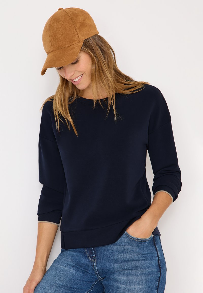 Marineblauer Sweatshirt mit Rundhalsausschnitt und Dreiviertelärmeln, kombiniert mit einer braunen Wildleder-Basecap, gestylt mit verwaschenen blauen Jeans.