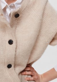 Béžový pletený cardigan s uvolněným střihem, který má ribbovanou strukturu, velké černé knoflíky a hluboký výstřih.