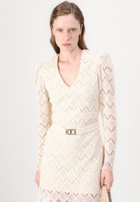 Mežģīņu kleita off-white krāsā ar V-veida kakla izgriezumu un garām piedurknēm. Piedāvā pieguļošu jostu ar zelta sprādzi un sarežģītu dimanta rakstu.