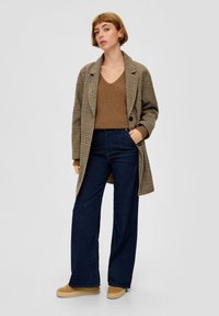 Manteau en laine à carreaux dans des tons de marron porté sur un pull en tricot marron, associé à un jean à jambes larges bleu marine et des chaussures à plateforme beiges.