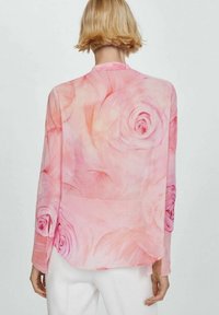 Blusa trasparente rosa con motivo a rose, maniche lunghe con dettagli a bottone e orlo curvo. Il tessuto sembra leggero e morbido.