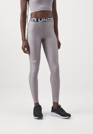 Donna che indossa leggings lilac Under Armour e un crop top abbinato, in piedi su uno sfondo bianco, con scarpe da ginnastica nere.
