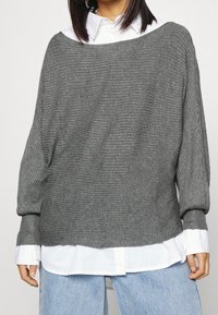 Grijze geribbelde trui met een brede halslijn en lange mouwen, gedragen over een wit overhemd met kraag, gecombineerd met lichtblauwe spijkerbroek.