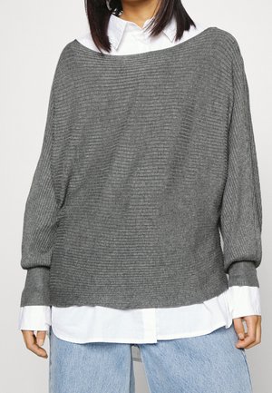 Grauer, gerippter Pullover mit weitem Ausschnitt und langen Ärmeln, darüber ein weißes Hemd mit Kragen getragen, kombiniert mit hellblauen Jeans.