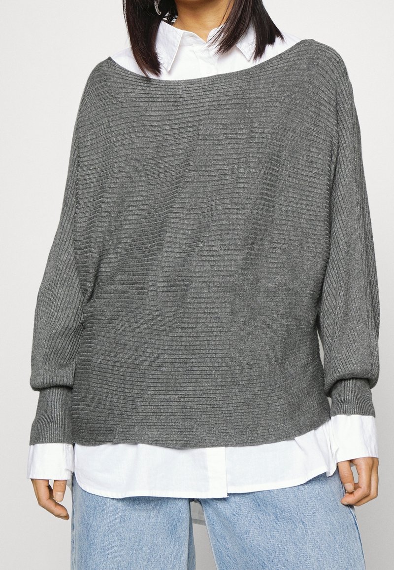 Grijze geribbelde trui met een brede halslijn en lange mouwen, gedragen over een wit overhemd met kraag, gecombineerd met lichtblauwe spijkerbroek.