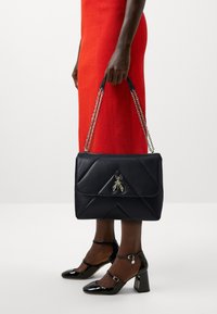 Borsa in pelle nera con design trapuntato, dotata di un emblema d'insetto dorato sulla parte anteriore. Accessori in catena sulle tracolle, sostenuta da una mano.