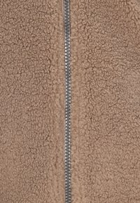 Tan fleecejakke med en tekstureret, blød overflade og en central lynlås. Lynlåsen har en sølvmetalisk finish for kontrast.