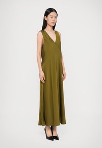 Hosszú olívazöld ruha mély V-nyakkivágással és ujjatlan kialakítással, oldalzsebekkel és könnyed, lengő sziluettel, fekete magassarkúval párosítva.