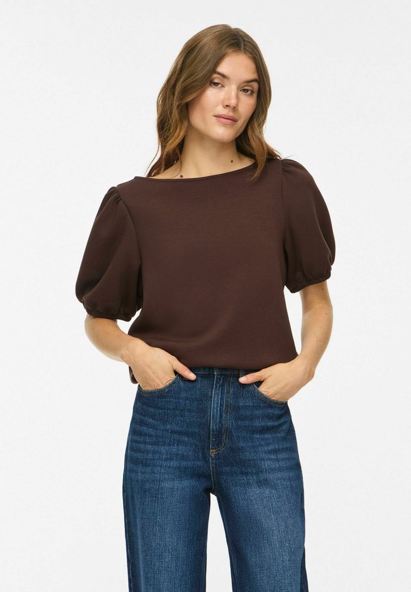 Femme portant un haut marron foncé à manches bouffantes et un jean bleu taille haute, debout les mains dans les poches sur un fond uni.