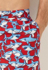 Bañador rojo con un patrón repetido de tiburones azules con acentos blancos, cinturilla elástica y bolsillo lateral para mayor comodidad.