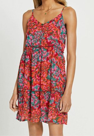 Vrouw draag een mouwloze bloemenjurk met een patroon van rood, roze, oranje en groen, gecombineerd met een fijn ketting en armbanden.