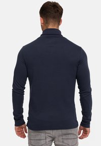 Homme portant une chemise bleu marine à manches longues et un pantalon gris, vu de dos sur un fond clair uni.