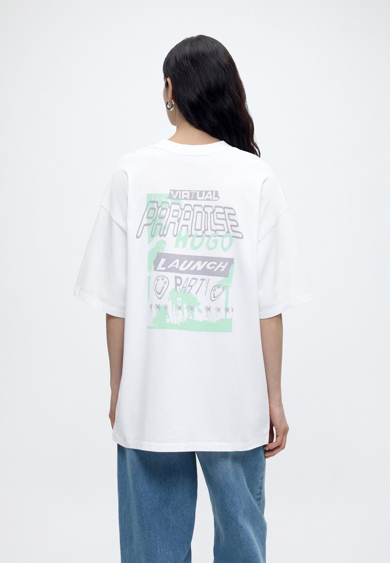 Personne aux longs cheveux foncés portant un T-shirt blanc oversize avec un texte graphique pastel et un motif de palmier dans le dos, associé à un jean bleu.