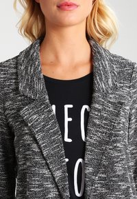Blazer texturizado en blanco y negro con solapas. La tela tiene un patrón tejido y cuenta con un diseño sin botones.