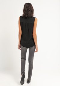 Femme aux cheveux mi-longs s'éloignant, portant un haut noir sans manches, un jean skinny gris et des chaussures noires sur un fond blanc.